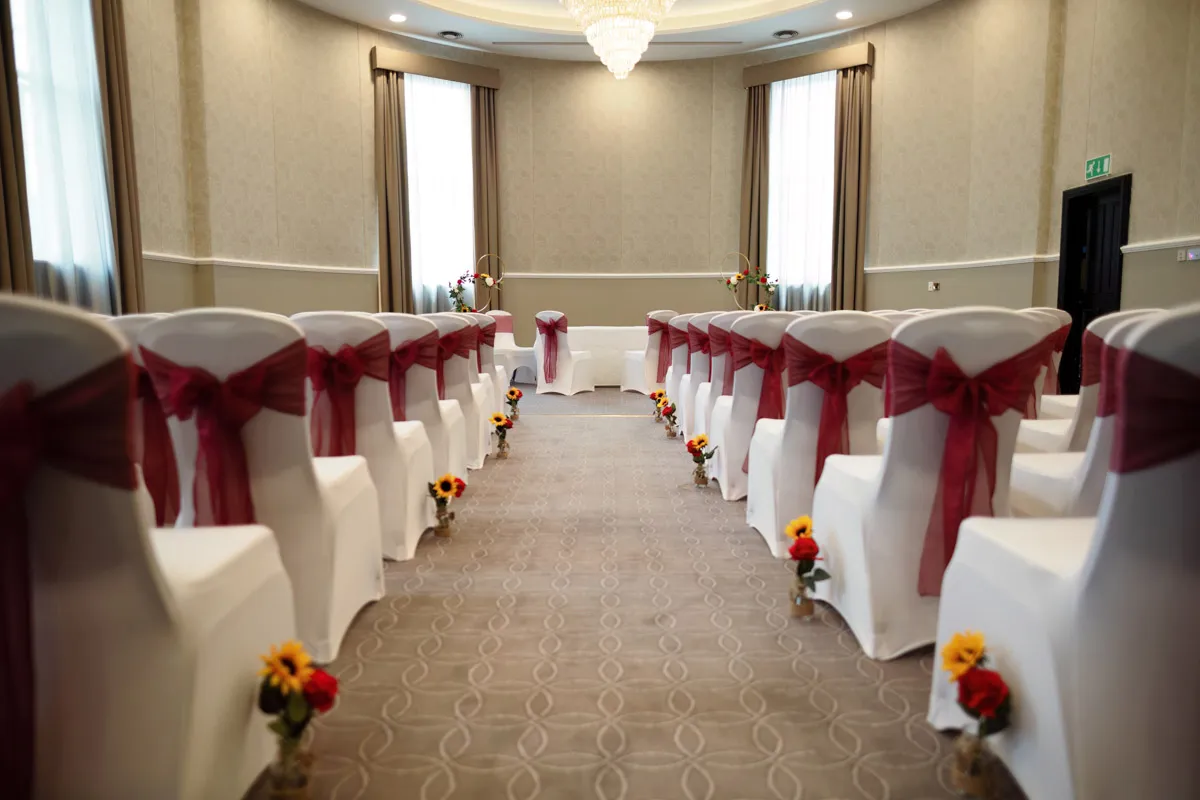 TJP_2023_06_24_0118 An elegant wedding ceremony setup inside a hall. White chair covers adorned with red sashes are arranged in two rows, facing an altar. The aisle is lined with small vases of colorful flowers, perfect for marrying your best friend. Large windows with curtains and a chandelier adorn the background.
