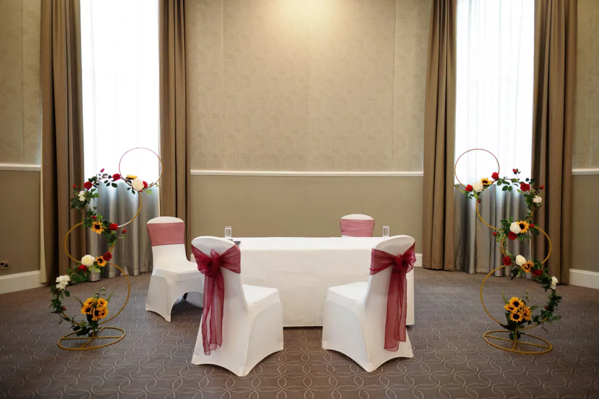 TJP_2023_06_24_0121 A decorated wedding ceremony space with two white chairs adorned with burgundy bows placed on each side of a white table. The room features beige walls, tall windows with sheer curtains, and circular floral arrangements with sunflowers and other colorful flowers, perfect for marrying your best friend.