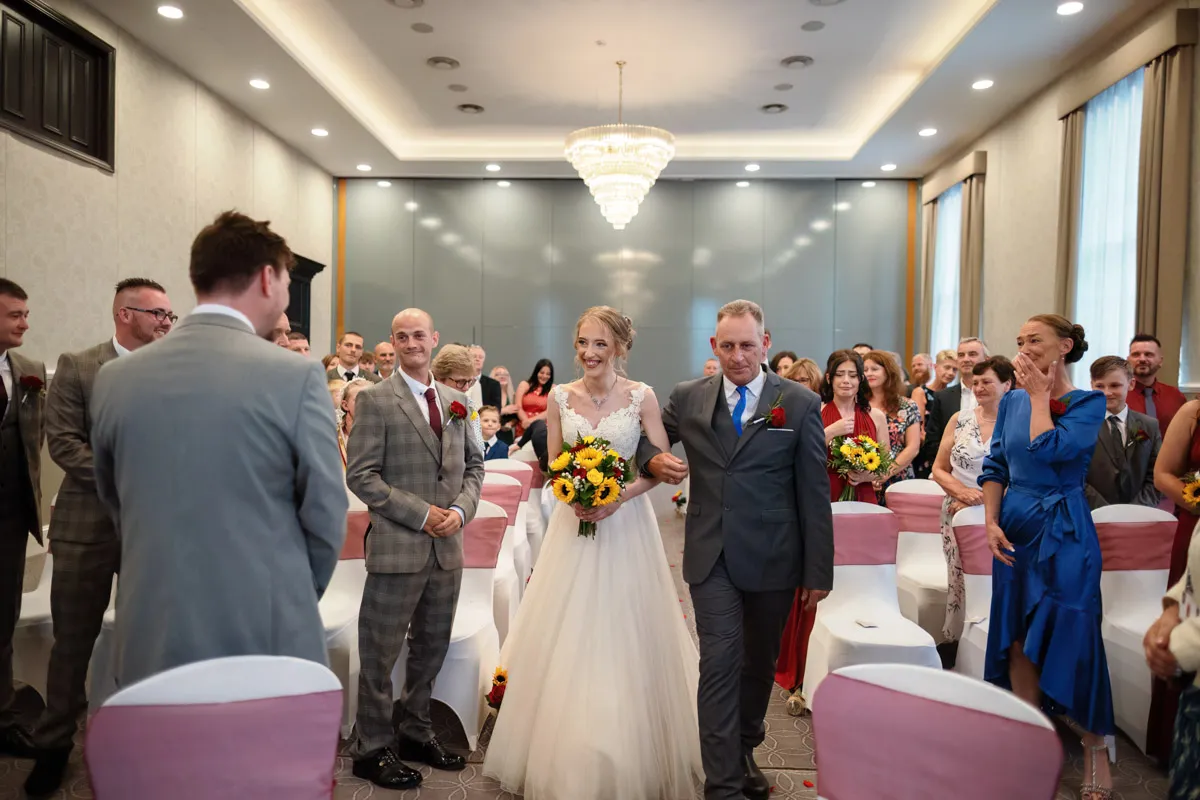 TJP_2023_06_24_0204 A bride in a white gown holding a bouquet of sunflowers walks down the aisle with her father, marrying her best friend in a heartfelt wedding ceremony. Guests are seated on either side, smiling and watching. The venue has white walls, large windows, and a chandelier overhead.