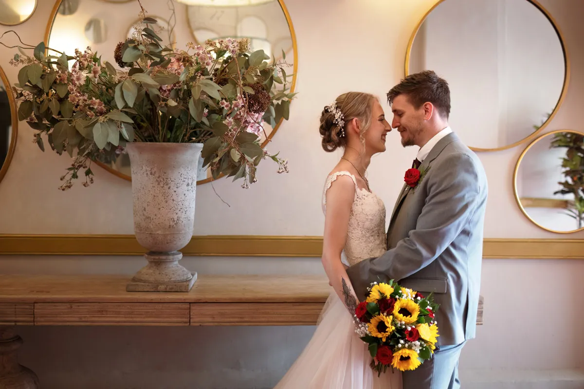 TJP_2023_06_24_0486 A bride and groom, marrying your best friend, stand close, gazing into each other's eyes in a warmly lit room. The bride holds a bouquet of sunflowers and red roses. Behind them is a tall vase with greenery and pink flowers, and round mirrors decorate the wall.