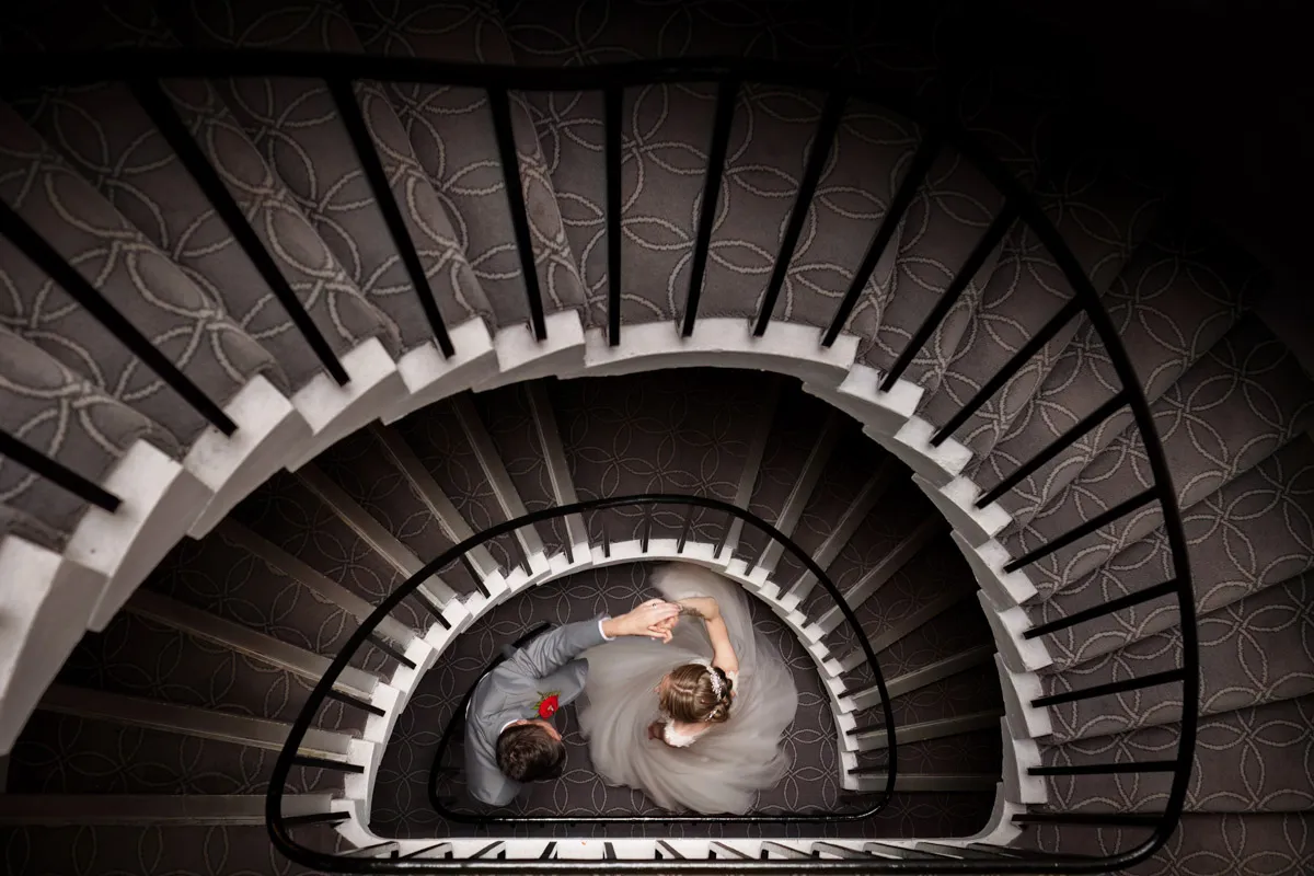 TJP_2023_06_24_0503 A bride and groom, embodying the essence of marrying your best friend, are seen from above holding hands at the bottom of a spiral staircase. The bride's white wedding dress contrasts beautifully with the groom's grey suit and red boutonniere. The staircase features dark railings and patterned carpet.