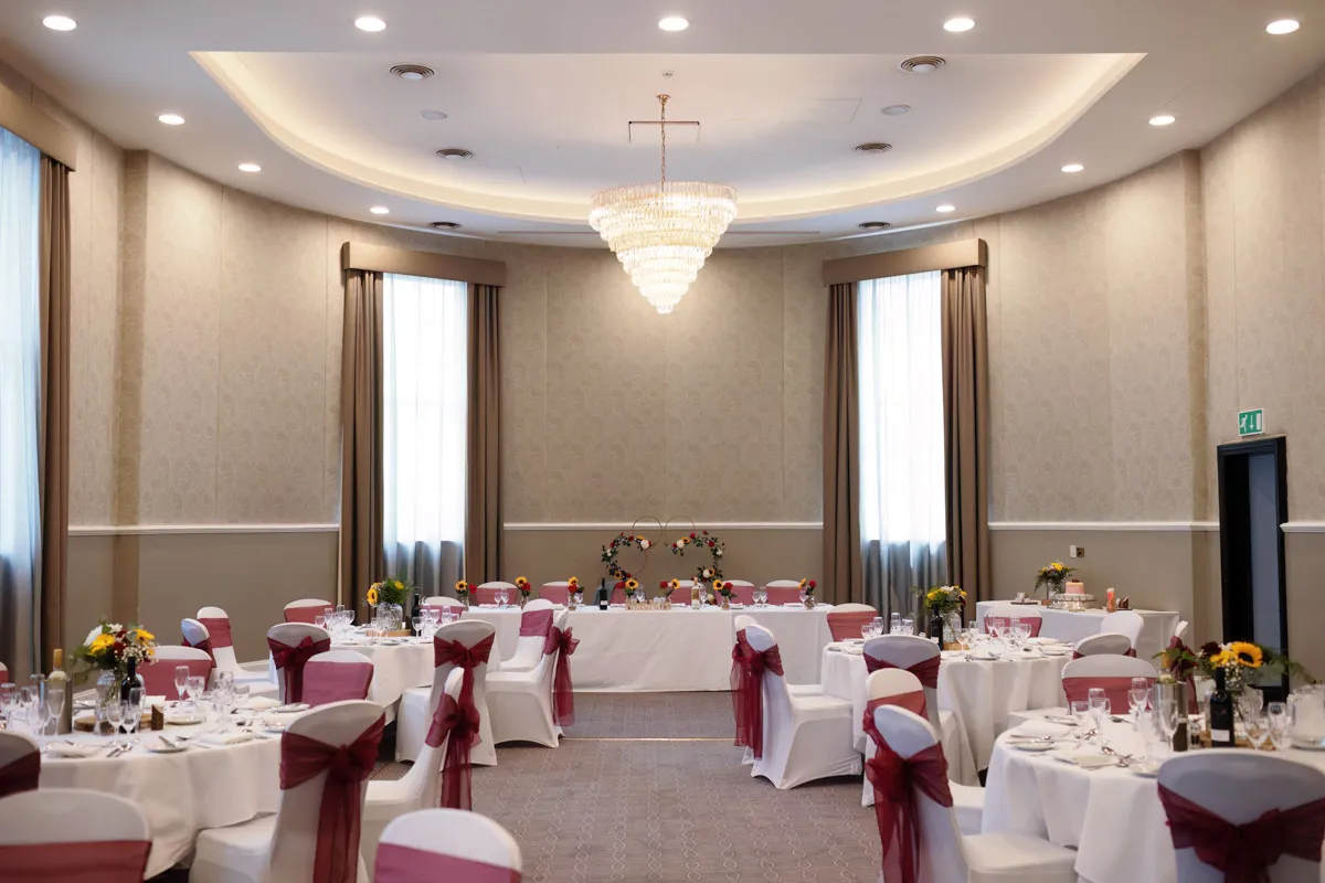 TJP_2023_06_24_0534 A large, elegantly decorated banquet hall with round tables dressed in white tablecloths and maroon sashes tied around the chairs. A long table at the front is adorned with floral arrangements perfect for celebrating marrying your best friend. A grand chandelier hangs from the ceiling, and windows with drapes line the walls.