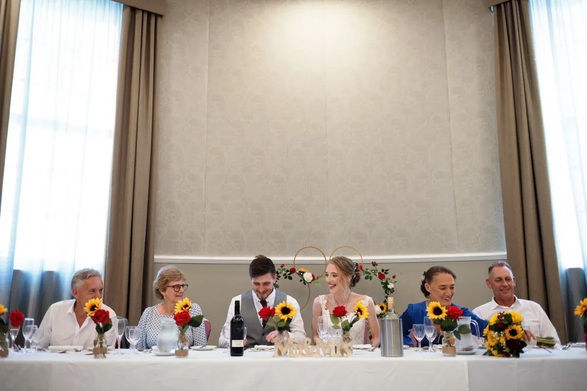 TJP_2023_06_24_0595 A long table at a wedding reception with six people seated. The couple, marrying your best friend, is centrally positioned, smiling in their wedding attire. The table is adorned with vibrant flower arrangements and wine glasses. Curtains hang behind them against a light-colored wall.
