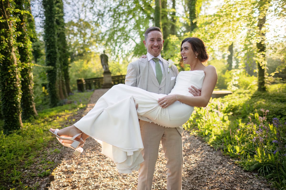 TJP_2024_05_05_0936 A groom in a light tan suit and green tie happily carries his laughing bride, who reflects the best bridal preparation, in a white dress through a sunlit forest path. Both are smiling, surrounded by lush greenery and dappled sunlight filtering through the trees.