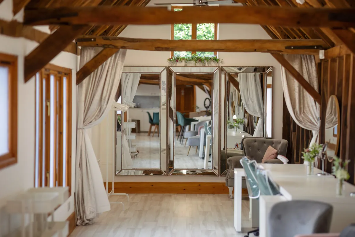 A stylish, light-filled salon with wooden beams and white interiors, perfect for yearly family portraits. The room features large mirrors, elegant white tables, and plush chairs. Soft curtains adorn the mirrors, and green plants add a touch of nature. In the background, more tables and chairs are visible.