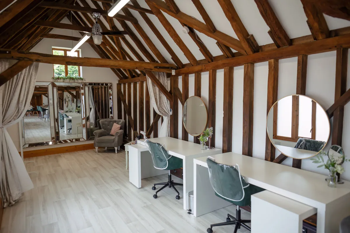 A chic, rustic interior featuring a room with white walls and wooden beams. Two white desks with minimalist chairs face circular mirrors. A cozy armchair is in the corner near a large mirror reflecting the room’s natural light from a window in the background—perfect for yearly family portraits.
