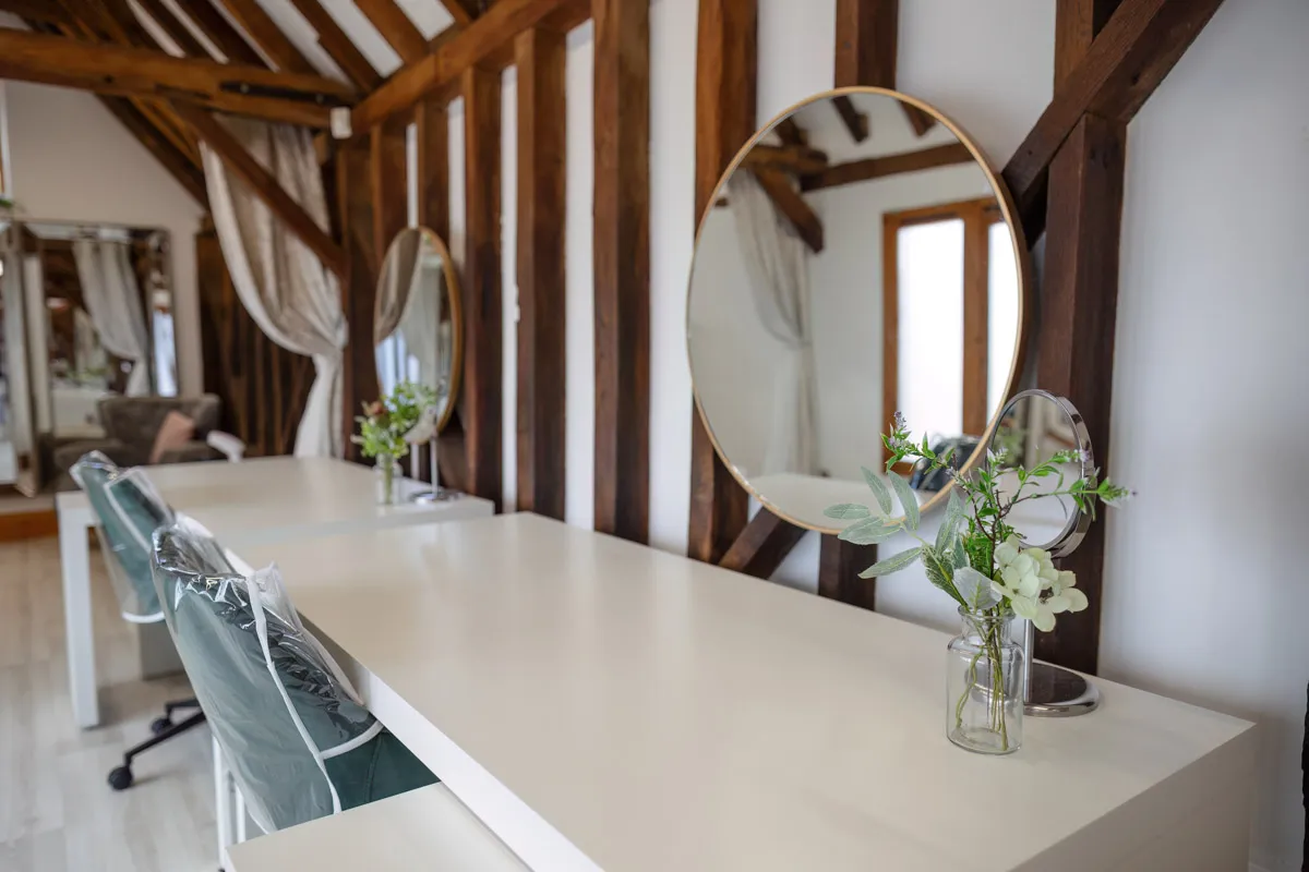 Interior of a modern salon with wooden beams and large round mirrors on white desks. Fresh flowers in clear vases and green chairs add to the decor, creating an inviting space perfect for yearly family portraits. Natural light from a window and reflective surfaces ensure excellent lighting throughout.