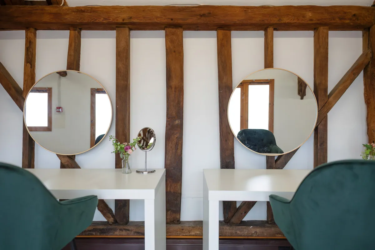 A room with rustic wooden beams adorns a wall behind two white desks. Each desk features a round mirror above it. On the left desk, there is a small vase with flowers and a smartphone, perfect for organizing yearly family portraits. Both desks are paired with green chairs, and ample natural light fills the space.