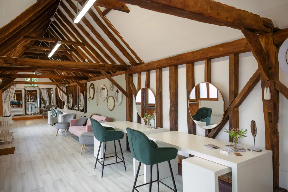 A stylish salon interior with exposed wooden beams and a vaulted ceiling. Modern seating, including green and pink chairs, line long white tables with mirrors. The light wood floor adds warmth to the space, perfect for capturing those yearly family portraits amidst plants and framed wall mirrors.