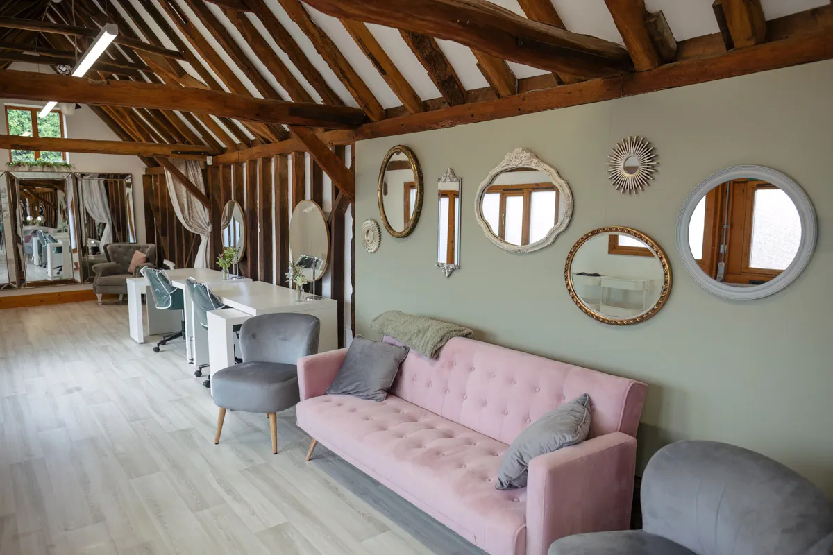 A stylish salon interior with wooden beams and light wooden flooring. The room features a pink sofa and two grey chairs against a light green wall adorned with multiple decorative mirrors, perfect for those yearly family portraits. In the background, white salon stations and chairs are visible.