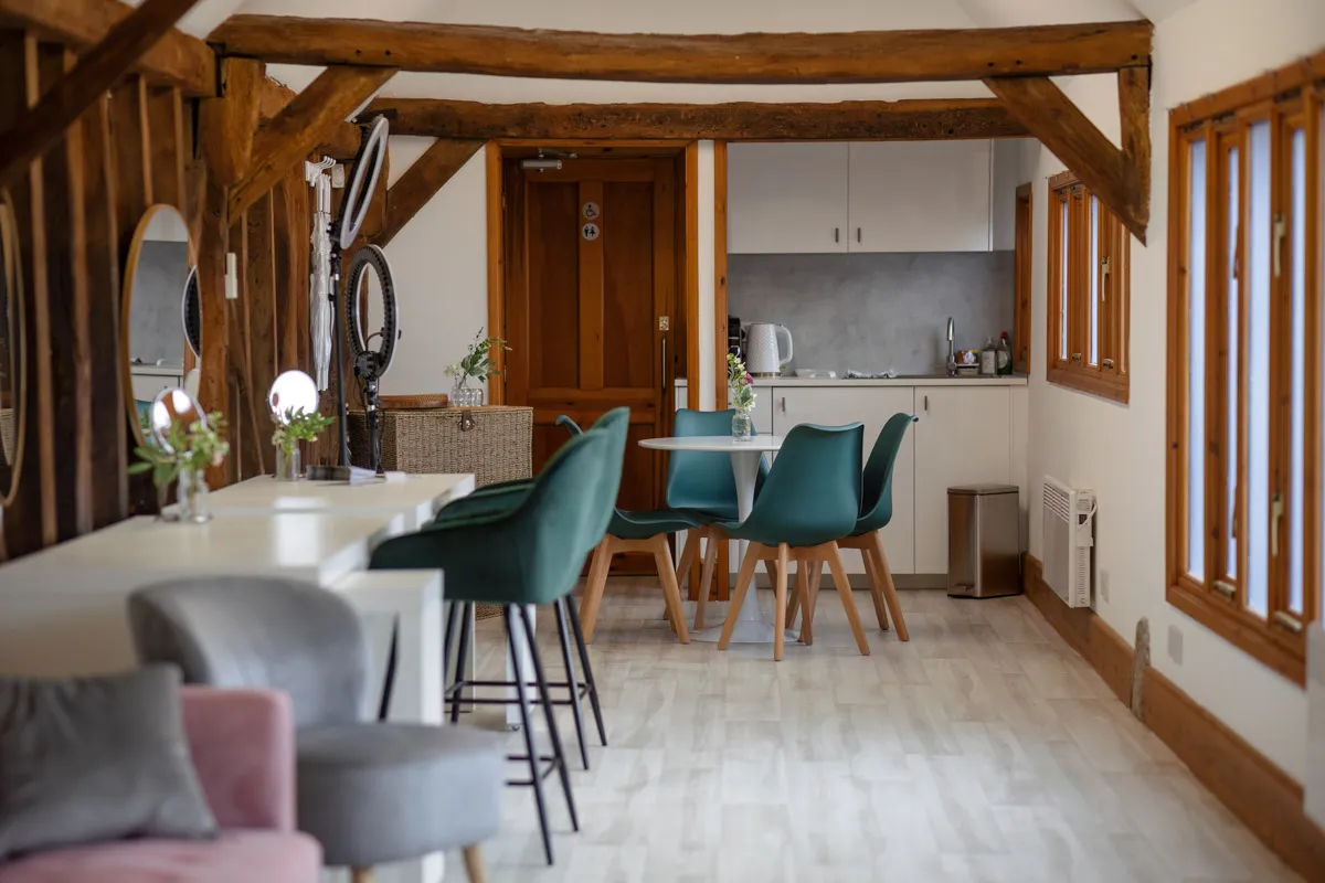 A cozy, rustic kitchen and dining area featuring exposed wooden beams. The space includes a small kitchenette with white cabinets, a dining table with green chairs, a seating area with a pink armchair, and a bar with stools. Natural light streams in through the windows, perfect for yearly family portraits.