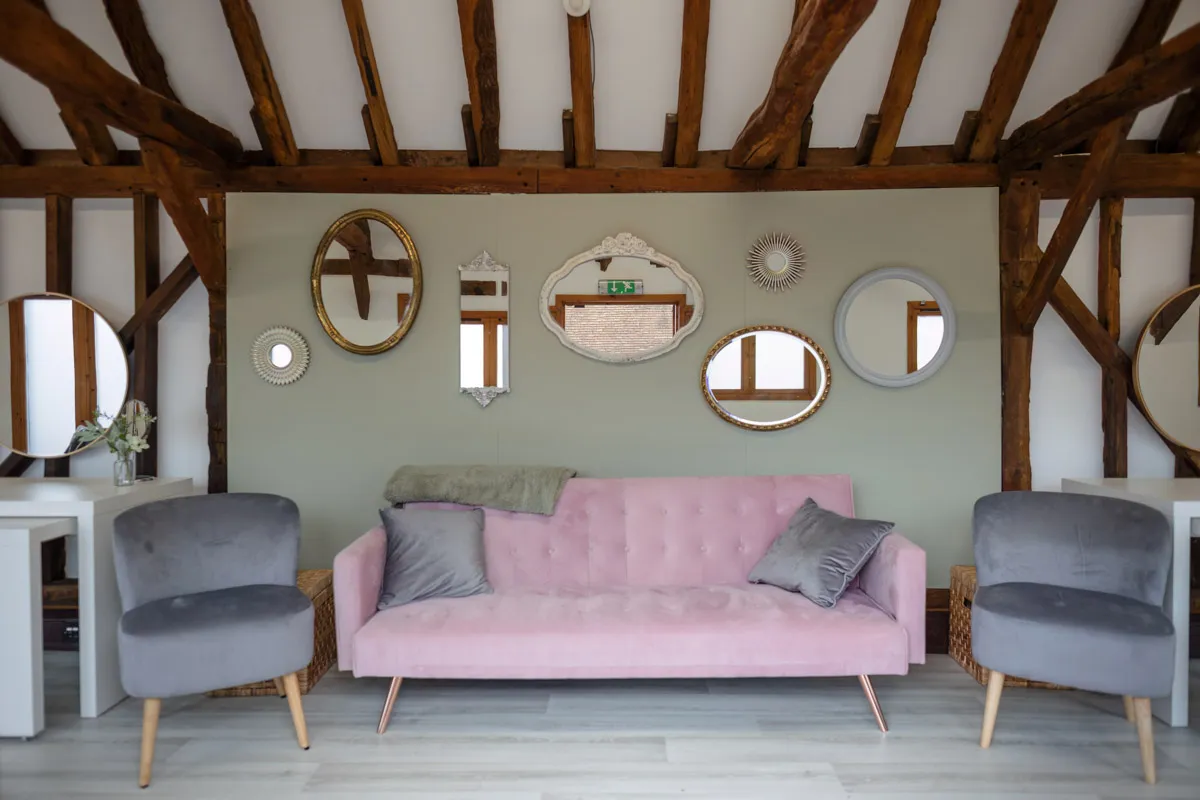 A cozy room with a vaulted wooden beam ceiling, featuring a pink tufted sofa adorned with gray pillows and a green throw. Flanked by two gray armchairs, the wall behind, often used for yearly family portraits, displays multiple decorative mirrors of various shapes and sizes.