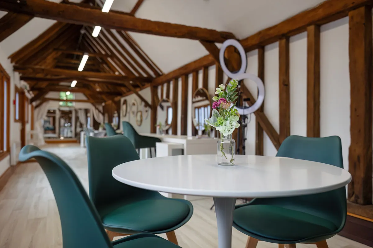 A modern office space with exposed wooden beams on the ceiling and walls. There is a round white table with green chairs around it, perfect for discussing projects or even displaying yearly family portraits. A small vase with flowers sits on the table. The room is well-lit with overhead lights and has a bright, airy atmosphere.