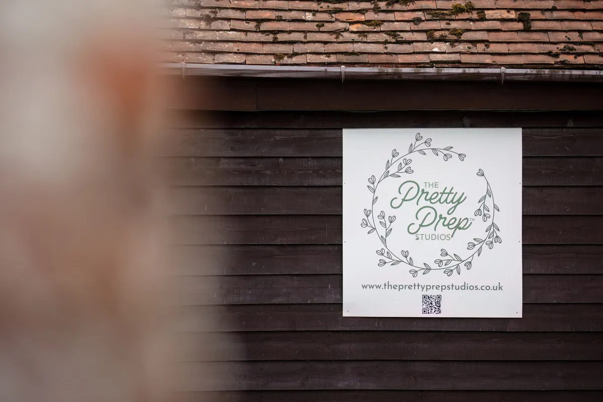 A wooden building with a white sign featuring the name "The Pretty Prep Studios" surrounded by a decorative leaf design. The sign mentions their yearly family portraits, along with a QR code at the bottom and the website URL www.theprettyprepstudios.co.uk. A blurred object is partially visible on the left.
