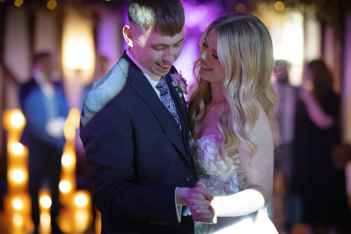 At the Villiers Hotel Buckingham wedding, a couple is dancing closely together, smiling and holding hands. The man is wearing a dark suit and the woman is in a light-colored dress. The background features warm, soft lights and blurred figures, creating a romantic atmosphere.