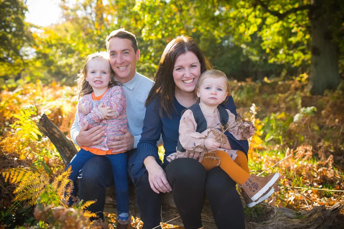 TJP_2017_10_27_00002 A skilled family photographer captures a family of four sitting on a log in a sunlit forest. The parents are smiling, holding their two young children amidst lush green foliage and scattered autumn leaves.