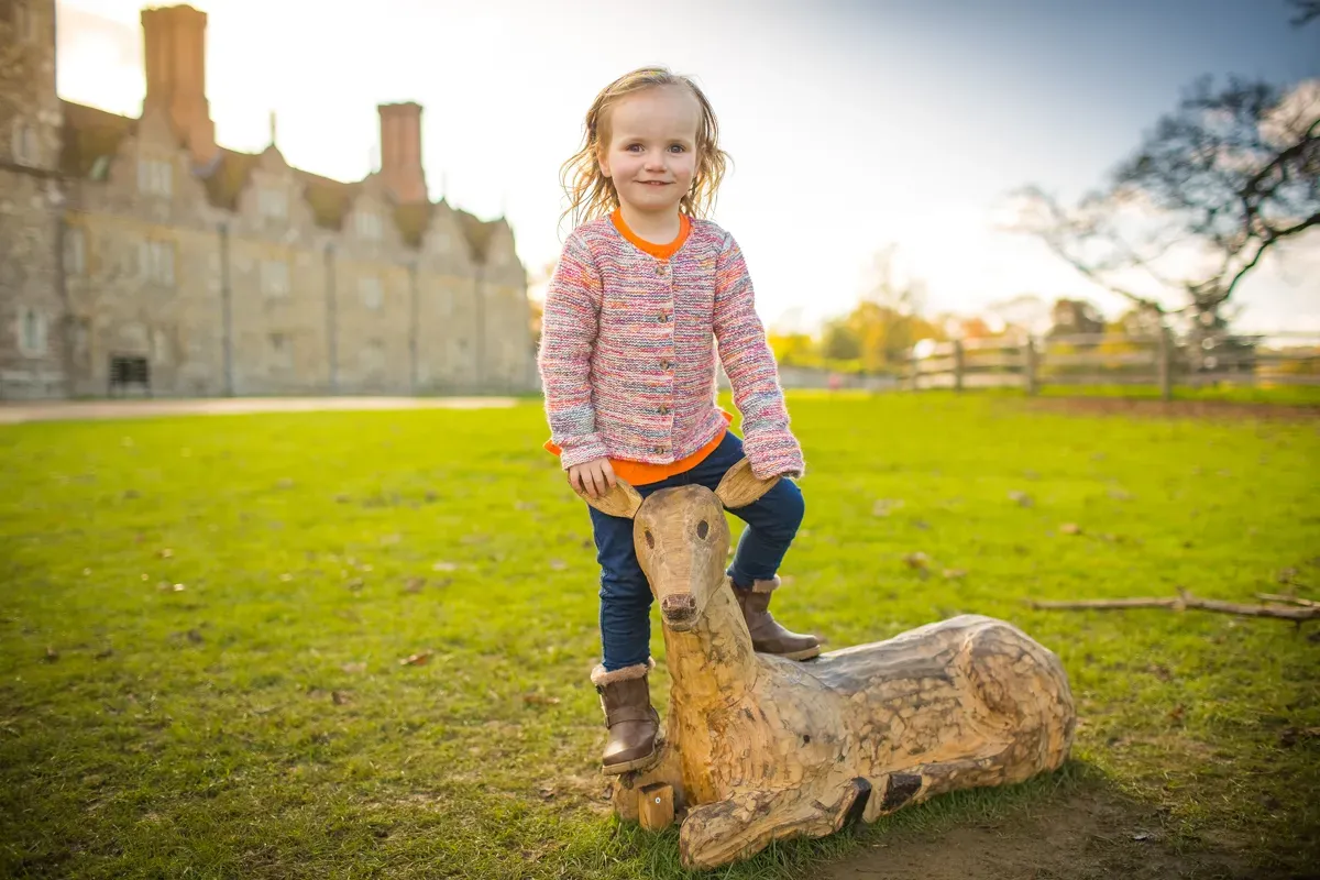 TJP_2017_10_27_00072 A young child in a colorful sweater and jeans poses playfully on a wooden deer sculpture amidst a grassy landscape. Captured through the lens of a family photographer, a historic stone building provides the perfect backdrop under a clear sky.