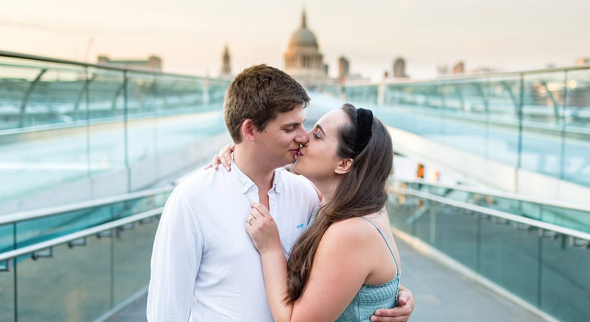 TJP_2020_08_09_0032_blog A couple shares a romantic kiss on a modern bridge at sunset, their joy captured flawlessly by Terence Joseph Photography. Surrounded by clear railings with a domed building in the backdrop, they are dressed casually, yet radiate happiness and love—an inspiring moment for wedding tips.