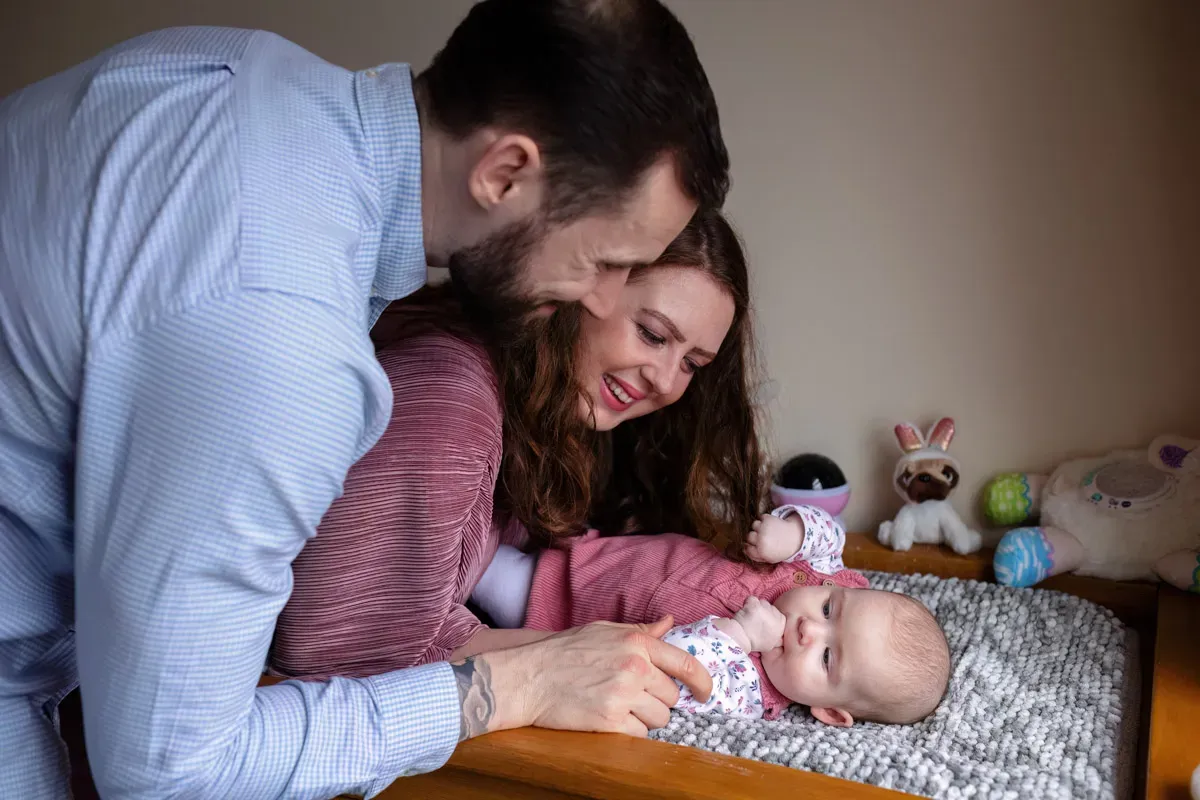 TJP_2024_02_09_0105 A smiling couple looks lovingly at their baby, who is lying on a changing table. Captured by a family photographer, the baby is in a patterned onesie with toys in the background. The scene is warm and intimate, lit gently by natural light.