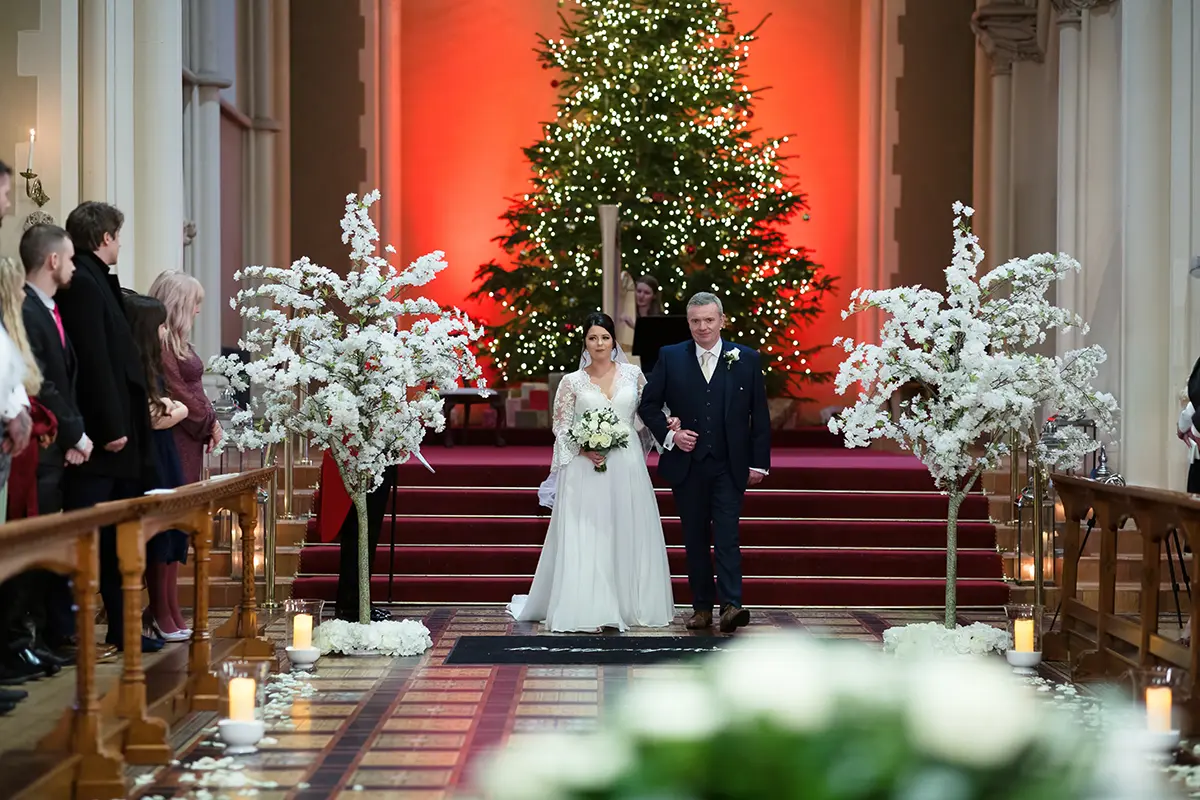 Blog_ How to Plan a Christmas Wedding A bride, holding a bouquet, walks down the aisle with a man in a dark suit. In their exquisite wedding venue near Buckingham, the hall is adorned with a large Christmas tree and glowing red lights. White floral arrangements line the aisle as guests watch this enchanting procession.