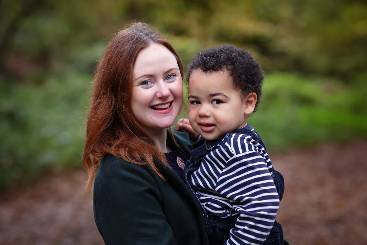 TJP_2024_11_02_0001 A woman with red hair smiles warmly while holding a young child in a striped shirt during their Tonbridge family photo session. They stand outdoors, surrounded by a blurred backdrop of greenery and fallen leaves, capturing a perfect moment of joy.