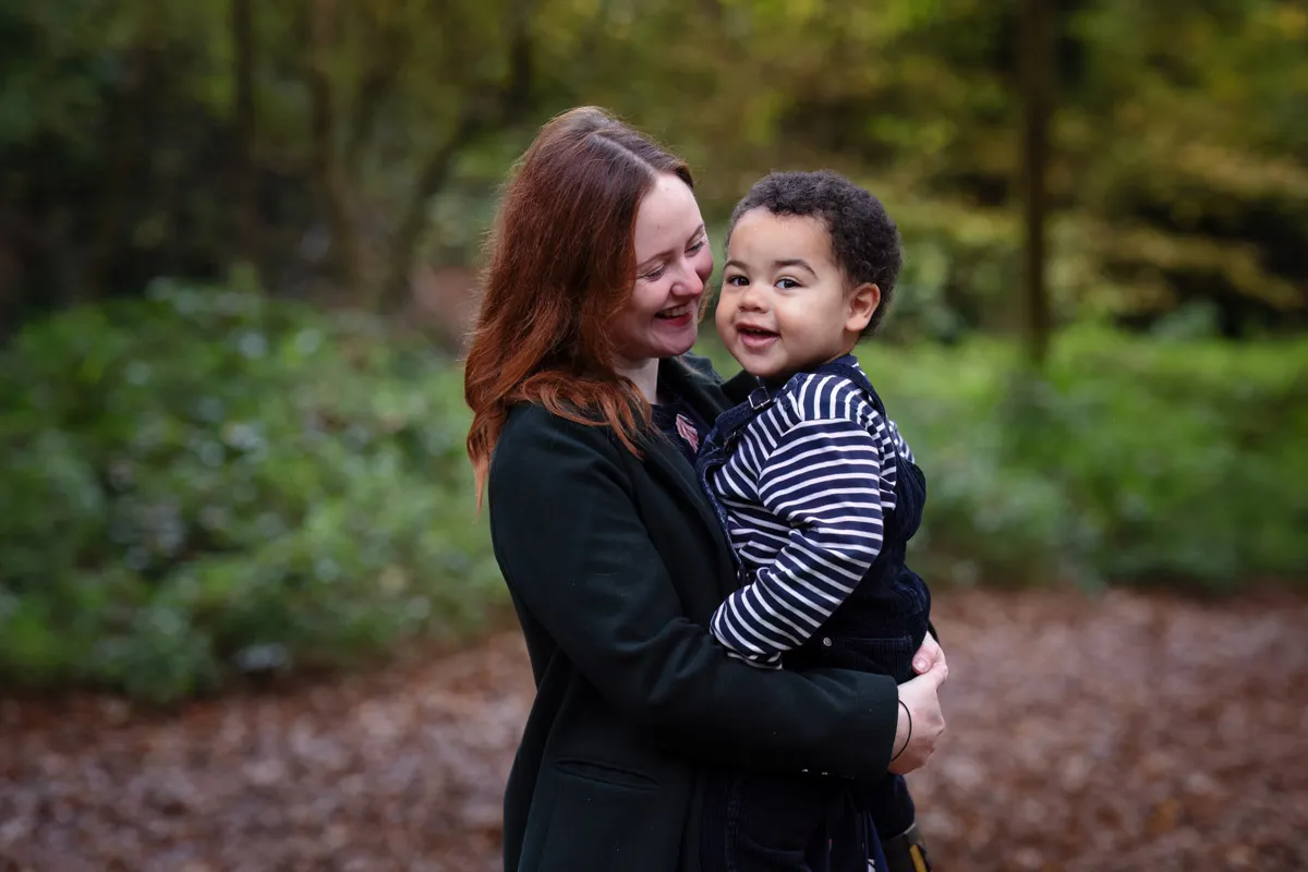 TJP_2024_11_02_0004 A woman with long hair and a dark coat is holding a smiling young child in a striped shirt during their Tonbridge Family Photo Session. They are outdoors in a leafy, green area, both appearing happy and engaged with each other.