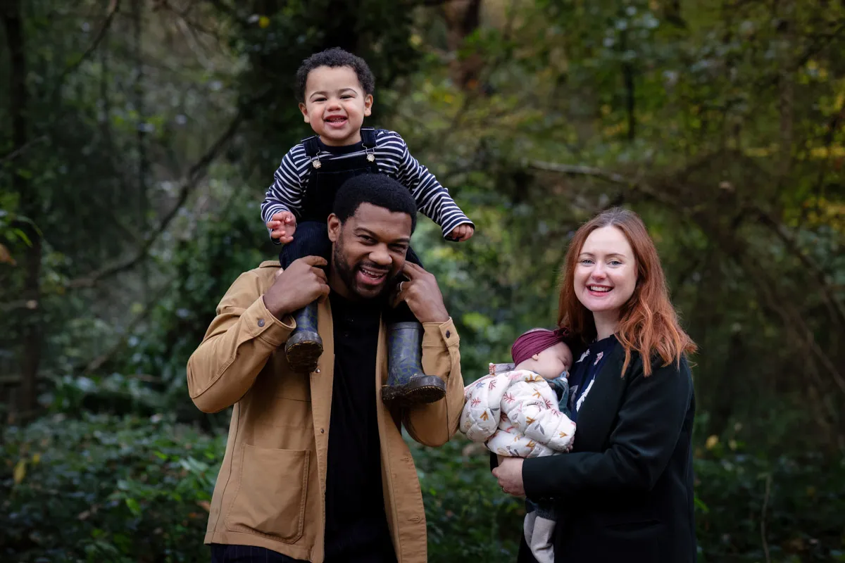 TJP_2024_11_02_0013 A smiling man stands in a wooded area with a young boy on his shoulders. Beside him, a woman with red hair holds a sleeping baby. All are dressed warmly and appear happy—captured perfectly by a Chislehurst Family Photographer.