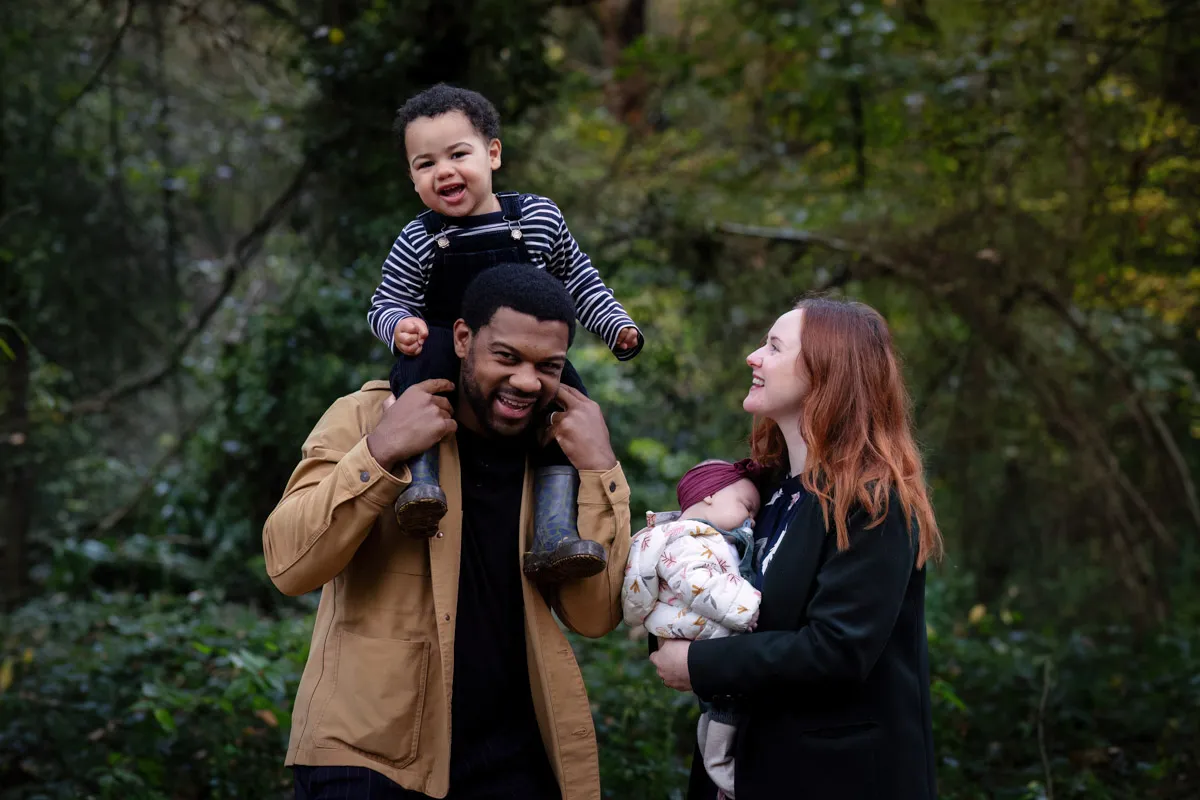 TJP_2024_11_02_0015 A joyful family delights in a Tonbridge Family Photo Session amid a lush forest. A man carries a laughing toddler on his shoulders, while a woman gently cradles a sleeping baby. The trees and greenery offer a serene backdrop for their happiness.