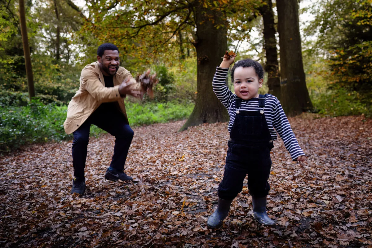TJP_2024_11_02_0019 In a delightful Tonbridge family photo session, a man and child joyfully play in an autumn forest. The man, donning a beige jacket, tosses leaves as the child, wearing striped overalls and boots, beams while holding up leaves. The ground is a carpet of brown foliage.