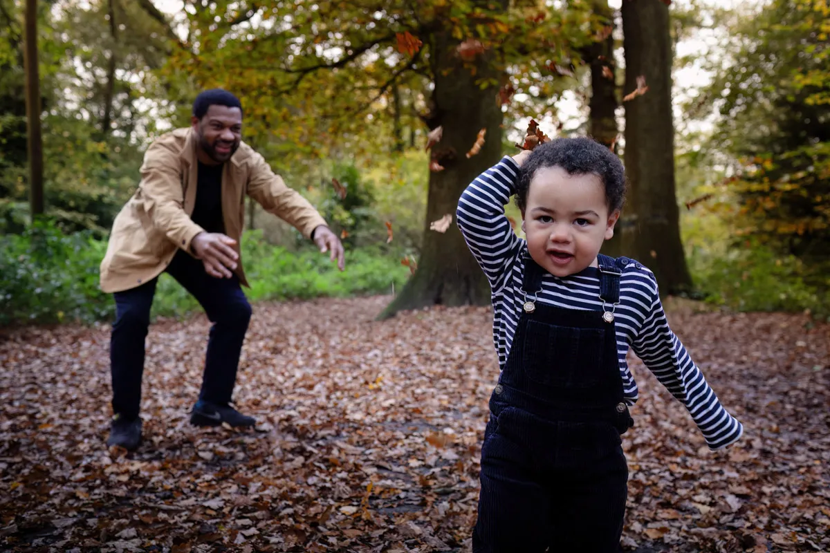 TJP_2024_11_02_0020 During a delightful Tonbridge Family Photo Session, a toddler in a striped shirt and overalls joyfully runs through a forest carpeted with fallen leaves. In the background, an adult in a tan jacket playfully follows, as the vibrant autumn foliage creates a picturesque scene.