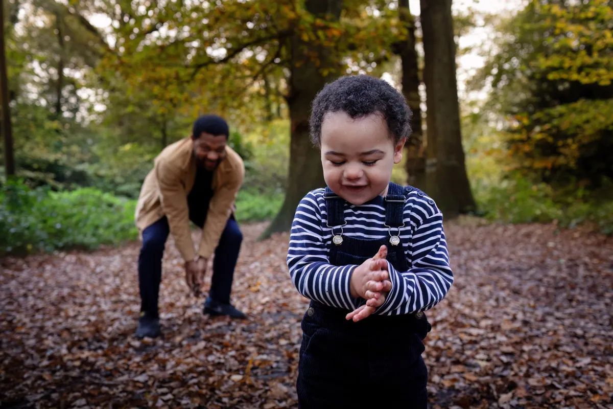 TJP_2024_11_02_0021 In a delightful Tonbridge Family Photo Session, a young child in striped overalls claps hands amidst fallen leaves in a forest. An adult in the background bends over, also surrounded by autumn foliage, capturing a playful and serene moment in nature.