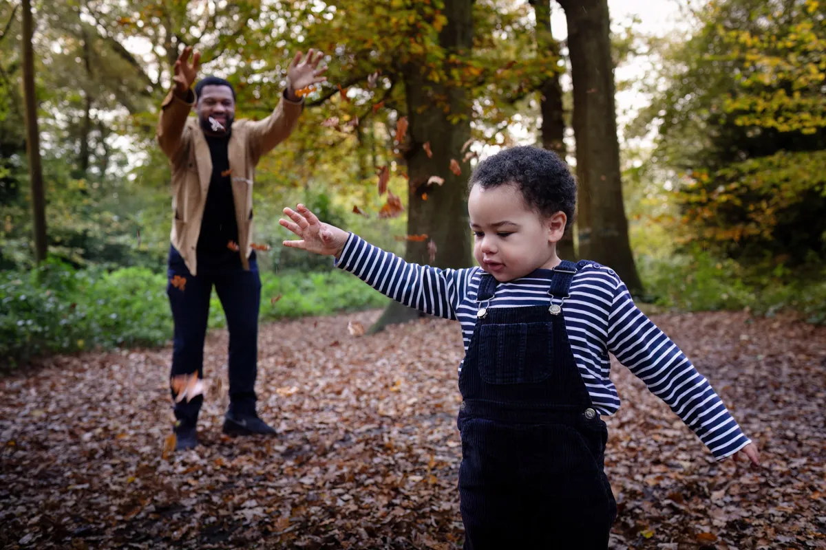 TJP_2024_11_02_0022 A child in a striped shirt and overalls plays in a forest during a Tonbridge family photo session, spreading their arms as autumn leaves fall. In the background, an adult joyfully tosses leaves into the air, surrounded by trees with vibrant foliage.