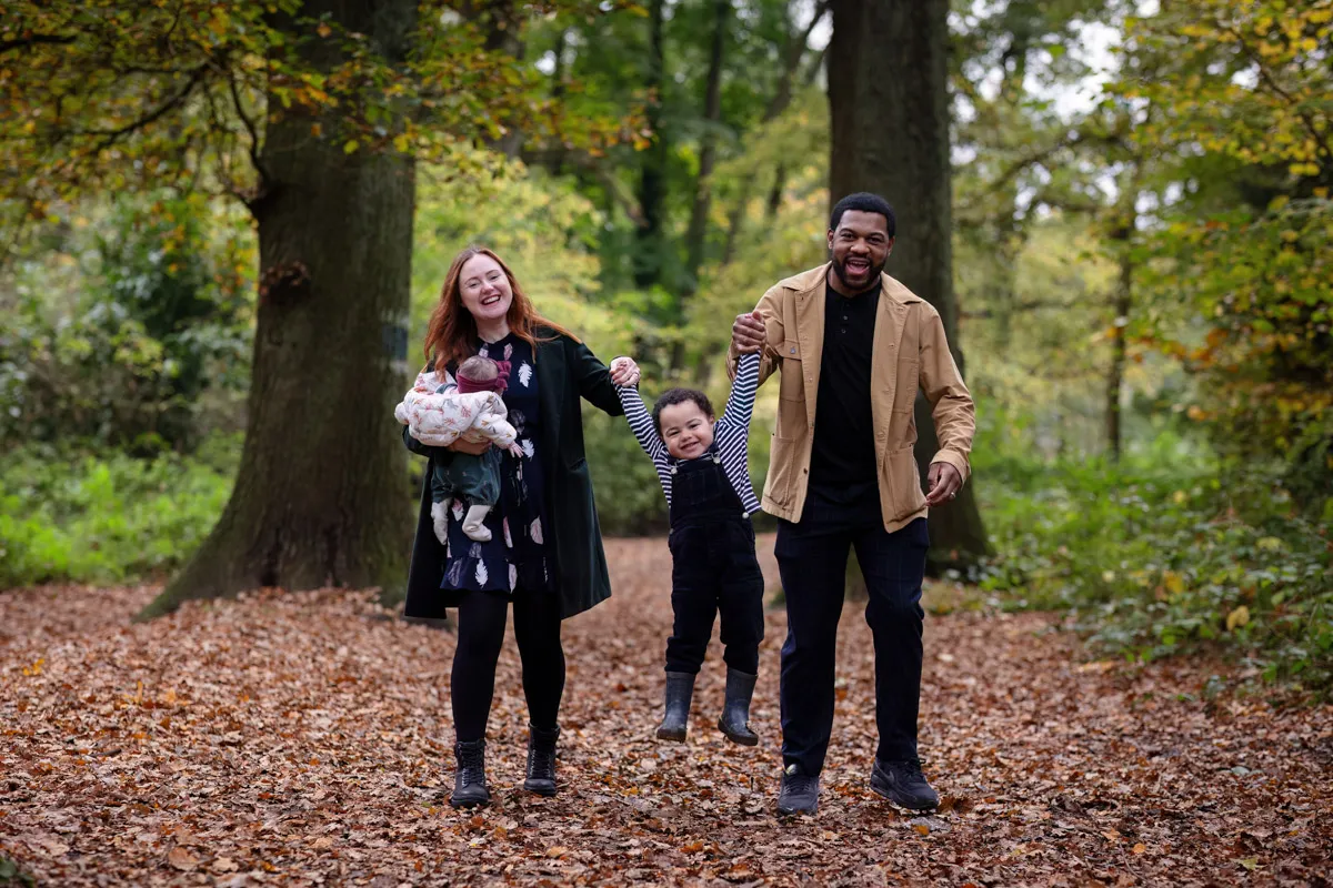 TJP_2024_11_02_0028 A family of four enjoys a walk in a forest during their Tonbridge Family Photo Session. The mother cradles a newborn, while the father swings a young child by the arms. They stroll along an autumn leaf-covered path, surrounded by tall trees. Everyone is smiling and dressed warmly.