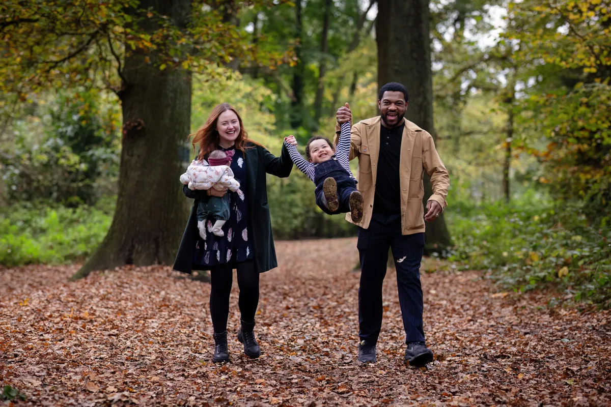 TJP_2024_11_02_0029 A joyful couple walks through a forest path covered in autumn leaves during their Tonbridge family photo session. The woman holds a baby, while the man swings a toddler by the hands, both smiling amid tall trees and lush foliage.