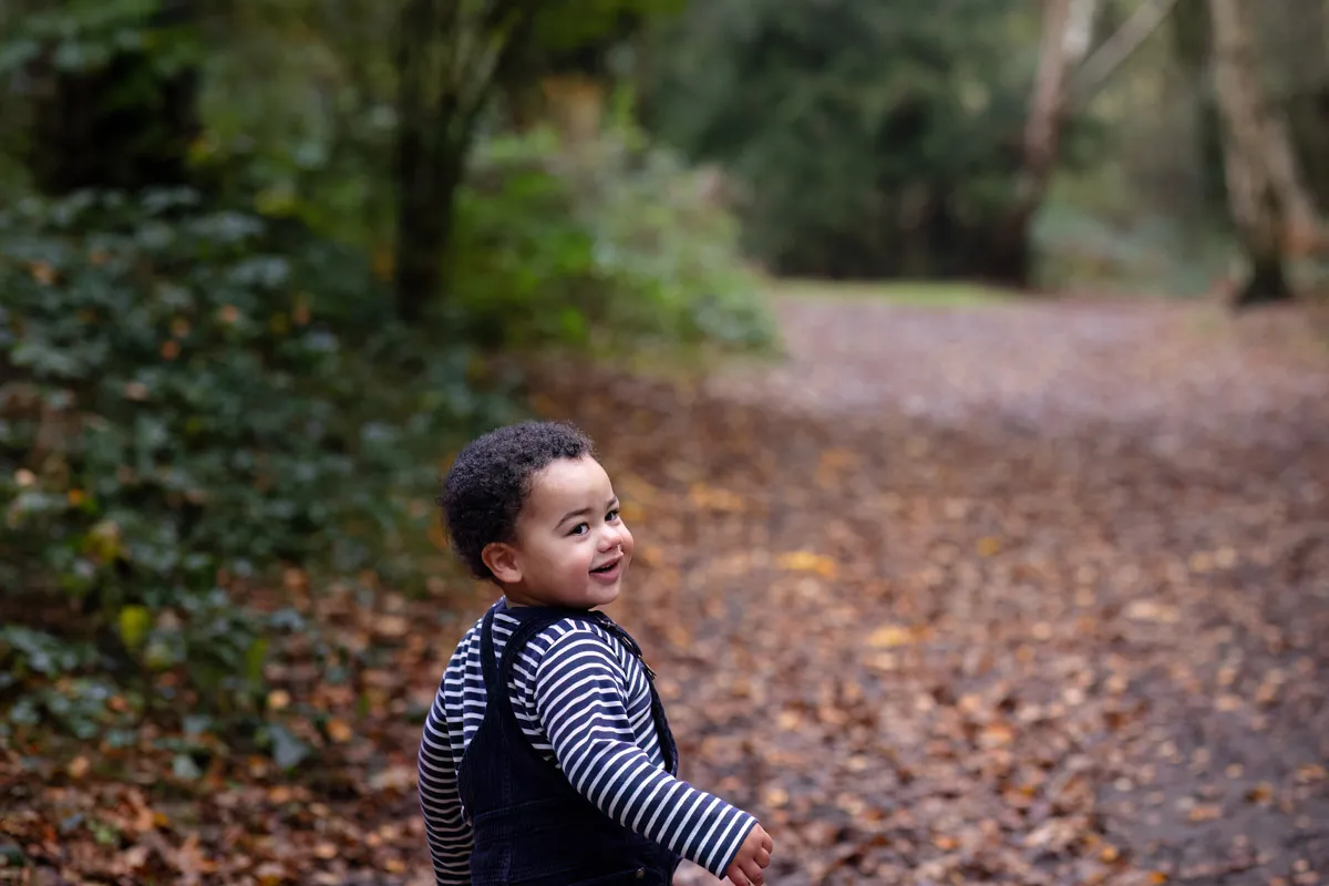 TJP_2024_11_02_0030 A young child with curly hair, wearing a striped shirt and dark overalls, is smiling as they walk along a leaf-covered path during a Tonbridge Family Photo Session. The trees are lush and green, capturing the essence of a vibrant autumn day in the forest.