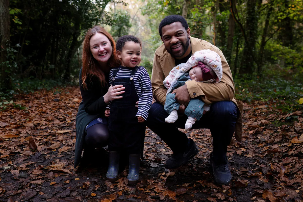 TJP_2024_11_02_0033 In this Tonbridge family photo session, a family of four poses in a forest. A woman kneels beside a young smiling child in overalls and boots. A man crouches holding a sleeping baby in a patterned hat. Leaves cover the ground, with trees in the background.