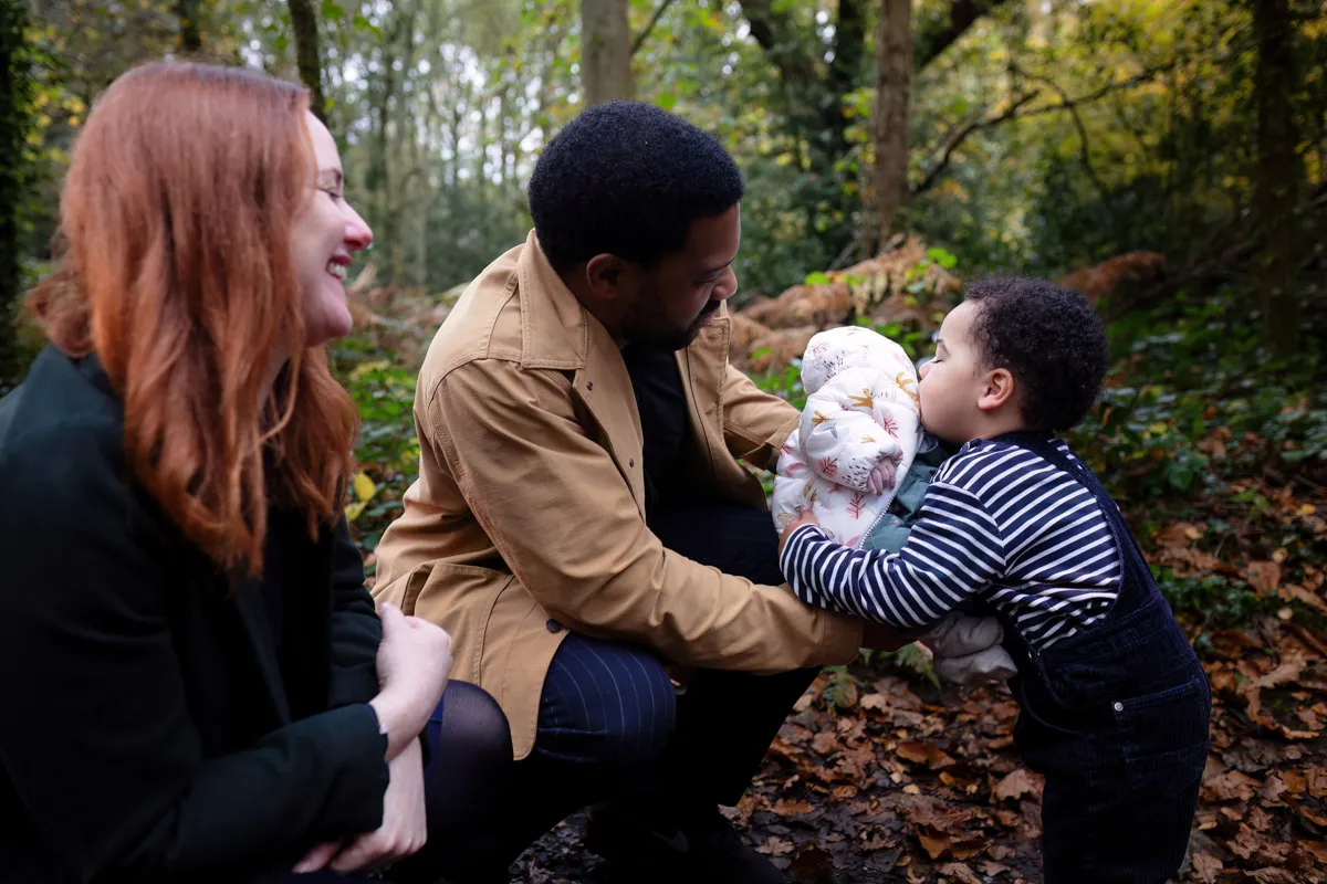 TJP_2024_11_02_0034 In this Tonbridge family photo session, a man crouches in a forested area, holding a baby wrapped in a floral blanket. A young child in a striped sweater interacts with the baby while a red-haired woman watches them, smiling. Leaves beautifully cover the ground around them.