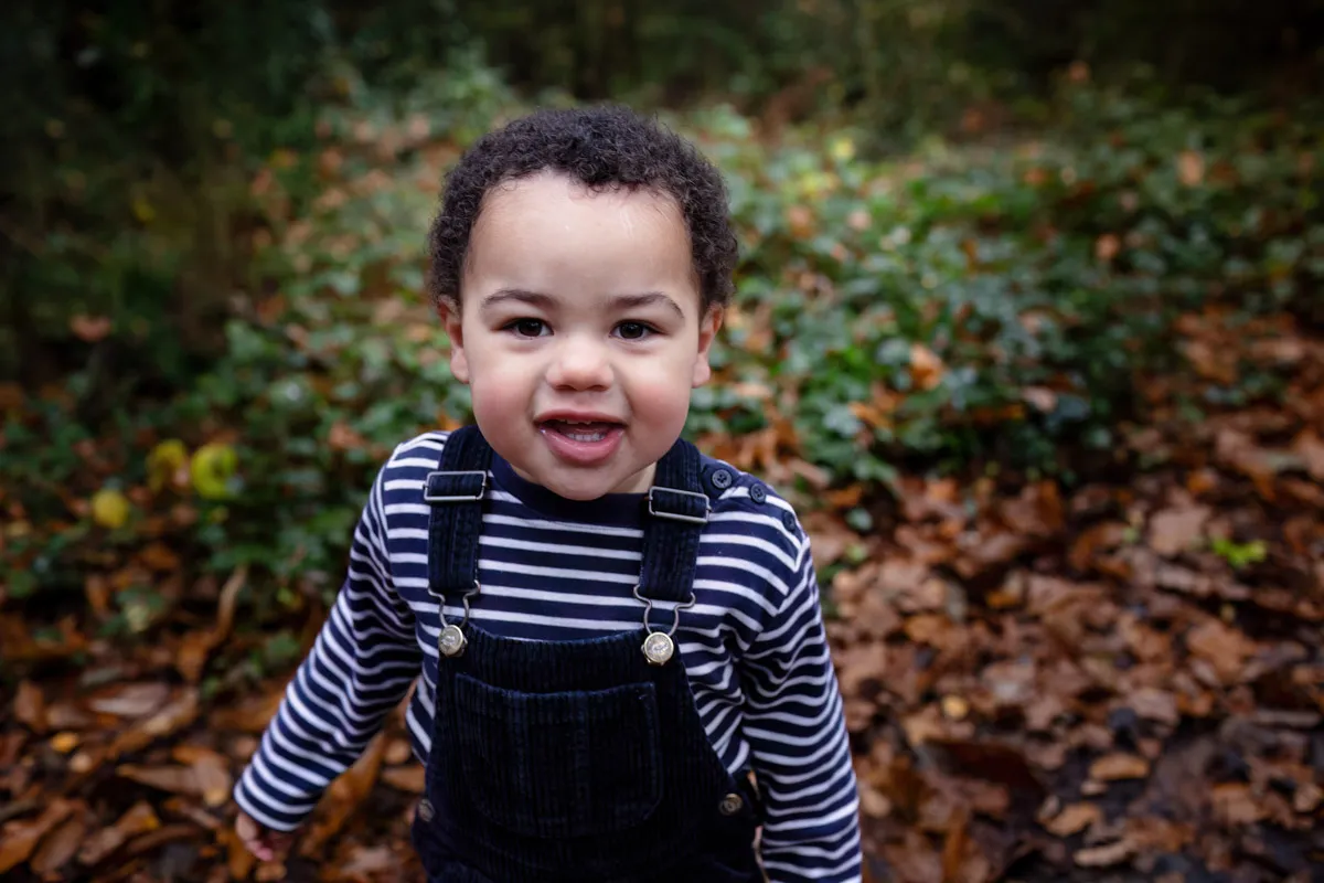 TJP_2024_11_02_0038 A young child in a striped shirt and overalls is smiling at the camera amidst an enchanting Tonbridge Family Photo Session. Standing outdoors, the ground is carpeted with autumn leaves, and lush green foliage provides a picturesque backdrop.