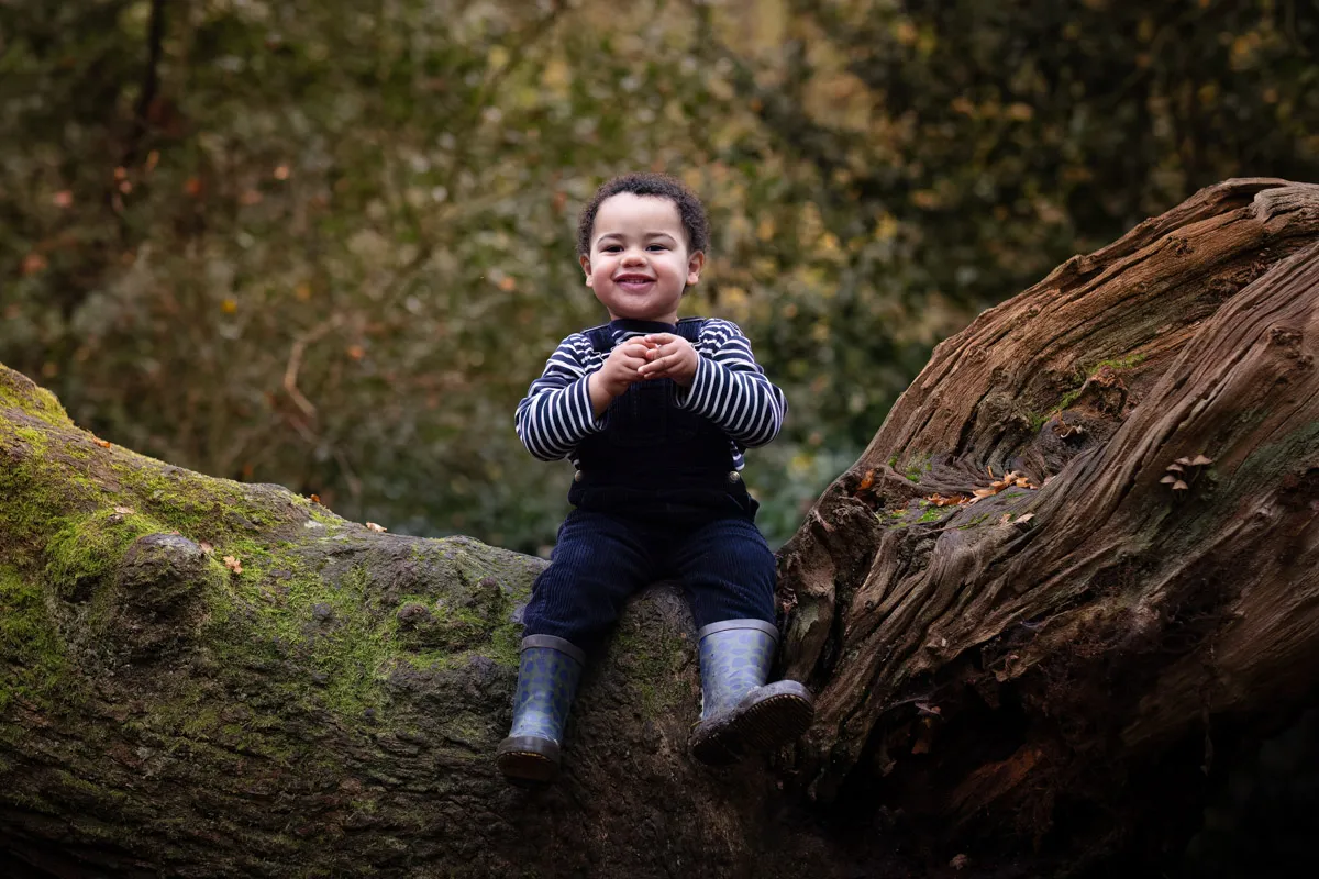 TJP_2024_11_02_0041 In a charming Tonbridge family photo session, a child in a striped shirt and overalls sits on a large, moss-covered tree trunk in the forest. They smile brightly in their rubber boots, surrounded by lush greenery.