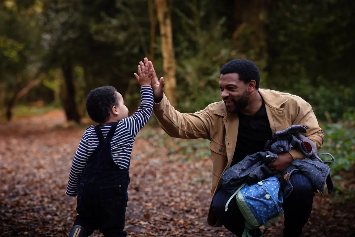 TJP_2024_11_02_0045 A man smiling as he kneels to high-five a young child in a forest during a Tonbridge family photo session. The man holds a backpack and jacket, while the child wears a striped shirt and overalls. They are surrounded by trees and fallen leaves on the ground, capturing the essence of their day.