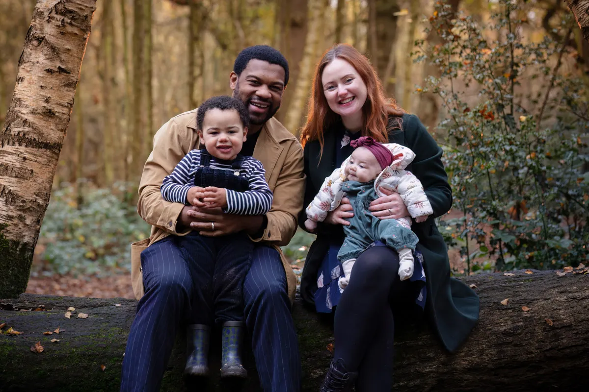 TJP_2024_11_02_0046 In a picturesque Tonbridge family photo session, a family of four sits on a log in the forest. The parents smile warmly as the father holds a young child in a striped shirt and the mother cradles a baby in a cozy jacket, while sunlight filters through the trees.