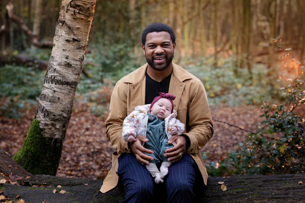 TJP_2024_11_02_0047 A smiling man sits on a fallen tree in a forest, holding a sleeping baby dressed in a floral jacket and maroon headband during their Tonbridge Family Photo Session. The background is filled with trees and autumn leaves covering the ground.