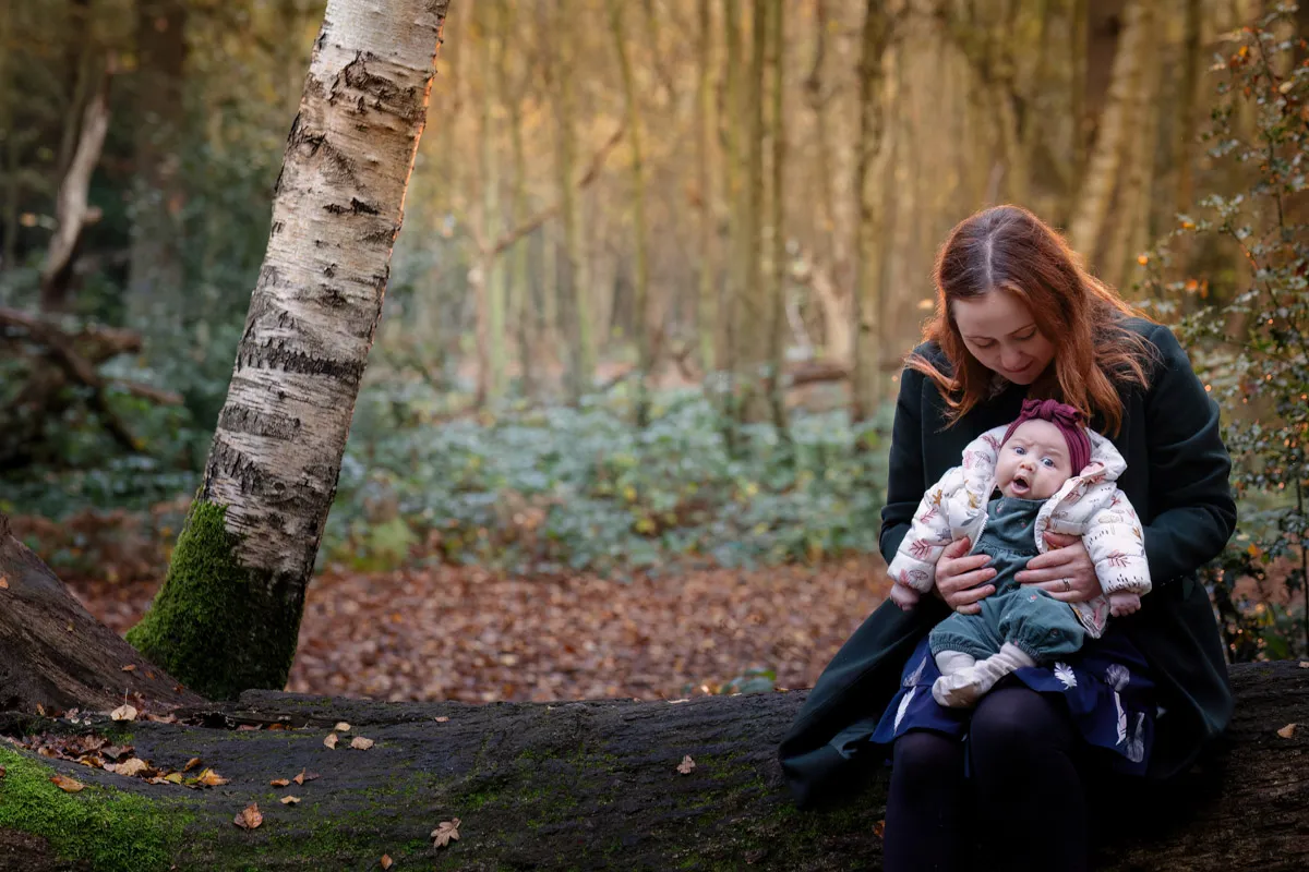 TJP_2024_11_02_0051 During a Tonbridge family photo session, a woman sits on a fallen tree trunk in the forest, holding her smiling baby. The baby is bundled in warm clothing and a hat, surrounded by tall trees adorned with autumn leaves.