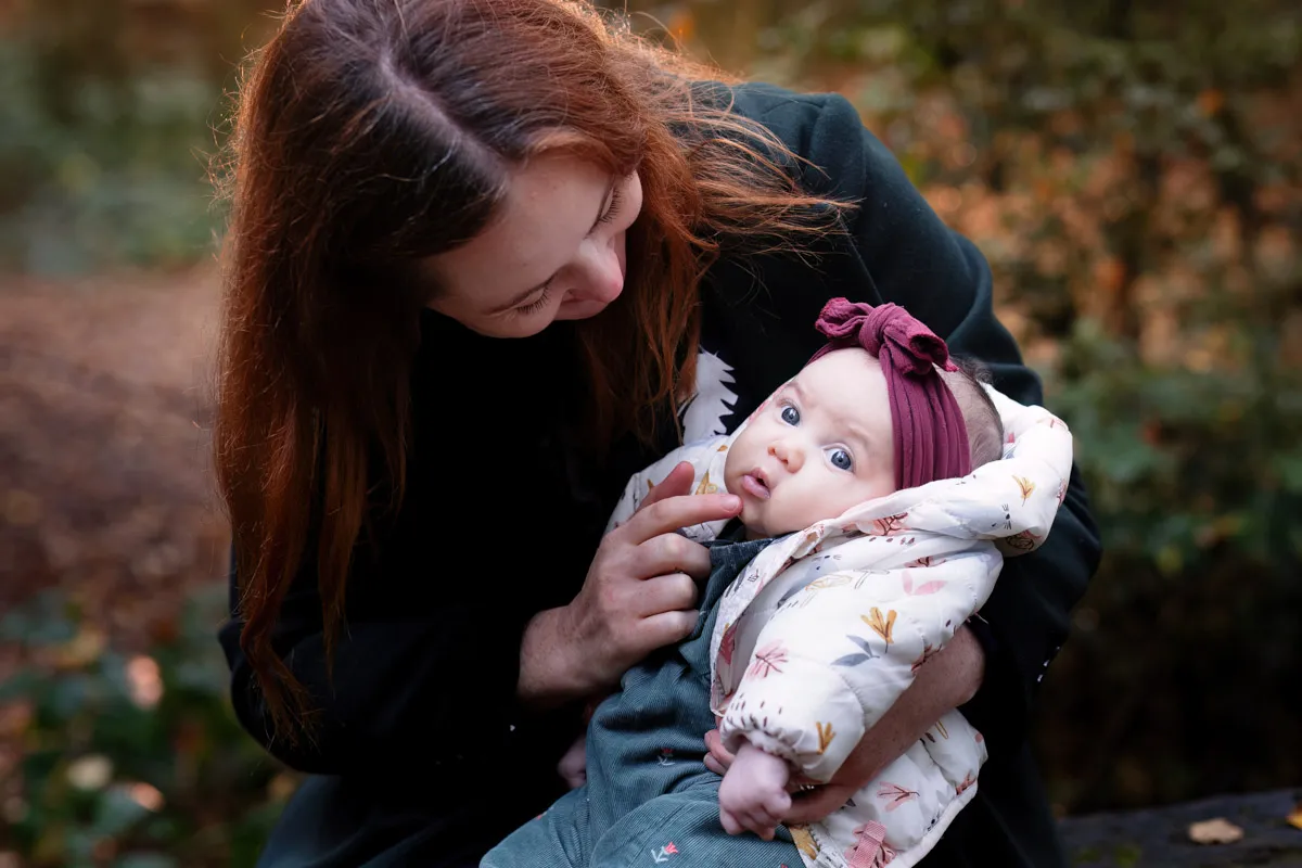 TJP_2024_11_02_0054 During a Tonbridge family photo session, a woman with long brown hair lovingly looks at a baby wrapped in a floral-patterned jacket and wearing a maroon headband. They are outdoors, surrounded by greenery and soft autumn lighting.