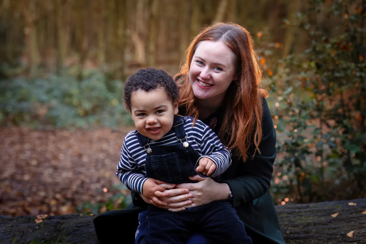 TJP_2024_11_02_0059 During a Tonbridge family photo session, a woman with long hair smiles while holding a young child in her lap. They are sitting outdoors, surrounded by trees and foliage. The child, wearing a striped shirt and overalls, looks happy and relaxed as the moment is beautifully captured.