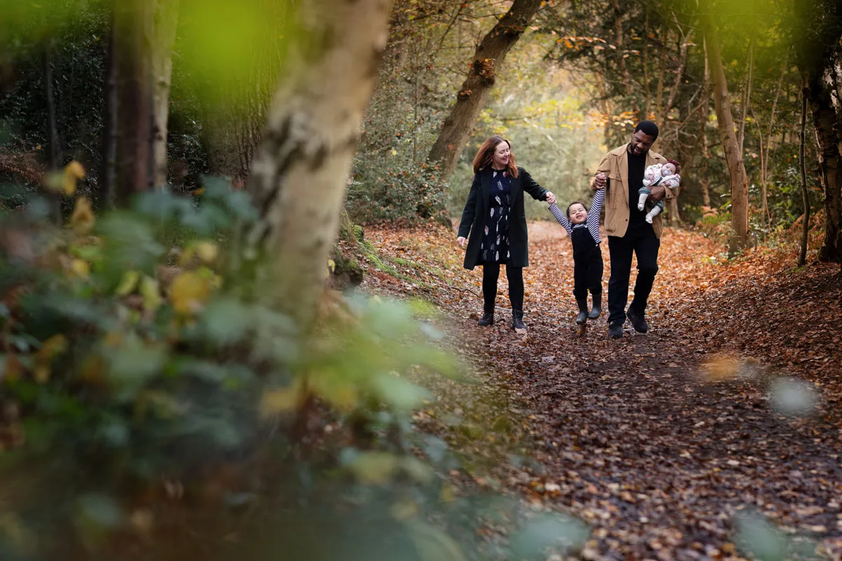 TJP_2024_11_02_0063 During their Tonbridge family photo session, a family of four strolls along a forest path blanketed in autumn leaves. The mother holds hands with a child, while the father cradles an infant. Sunlight filters through the trees, creating a serene atmosphere.