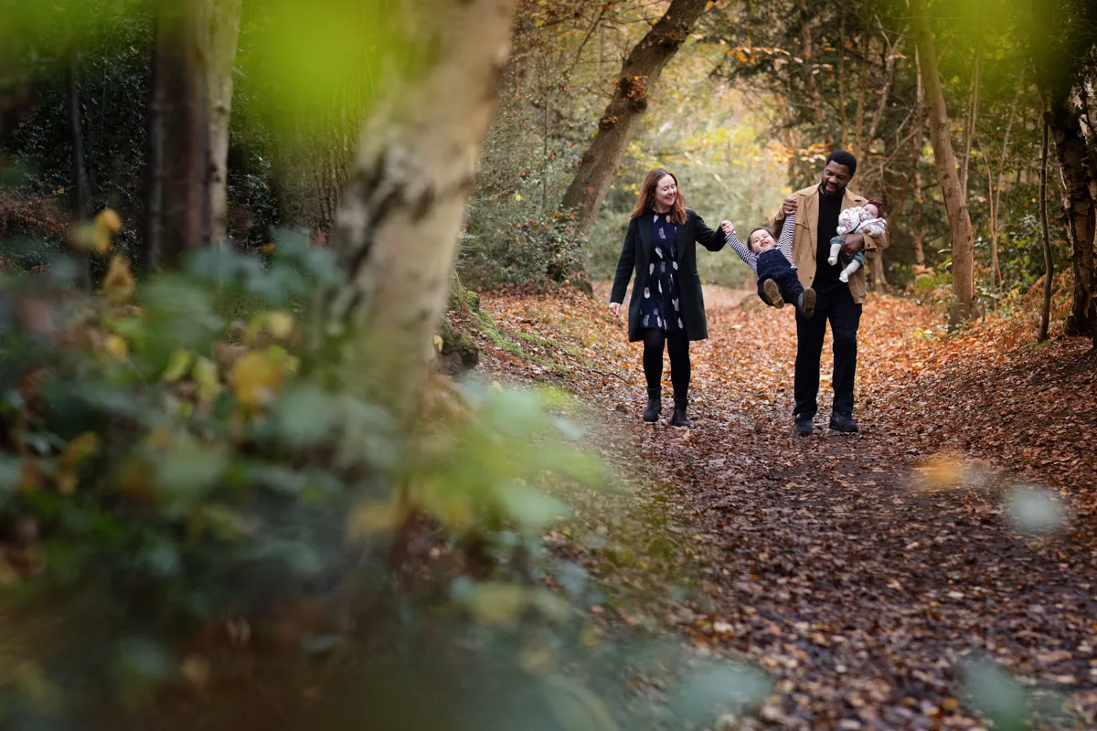 TJP_2024_11_02_0064 A couple enjoys a Tonbridge family photo session, walking along a forest path in autumn, smiling and holding hands with two small children. The path is covered with fallen leaves, and the sun filters through the trees, creating a warm, serene atmosphere.
