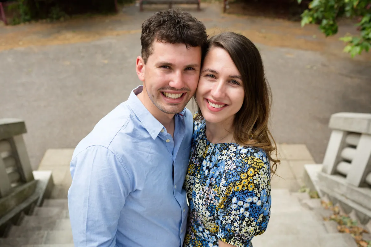 A smiling man and woman stand closely on stone steps, celebrating their cherry blossom engagement. The man wears a light blue shirt, and the woman dons a floral dress. They are outdoors with trees in the background, enjoying a sunny day.