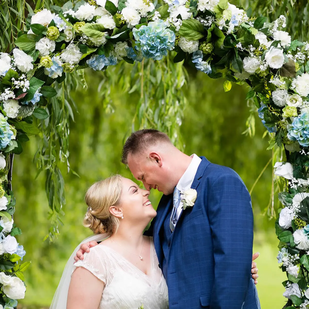 02_Liberty_Thomas-1_Creative_Wedding_Photographer A bride and groom stand smiling under a floral arch outdoors, touching foreheads lovingly. Captured in a creative wedding photography style, the arch is adorned with white and blue flowers and lush green leaves.