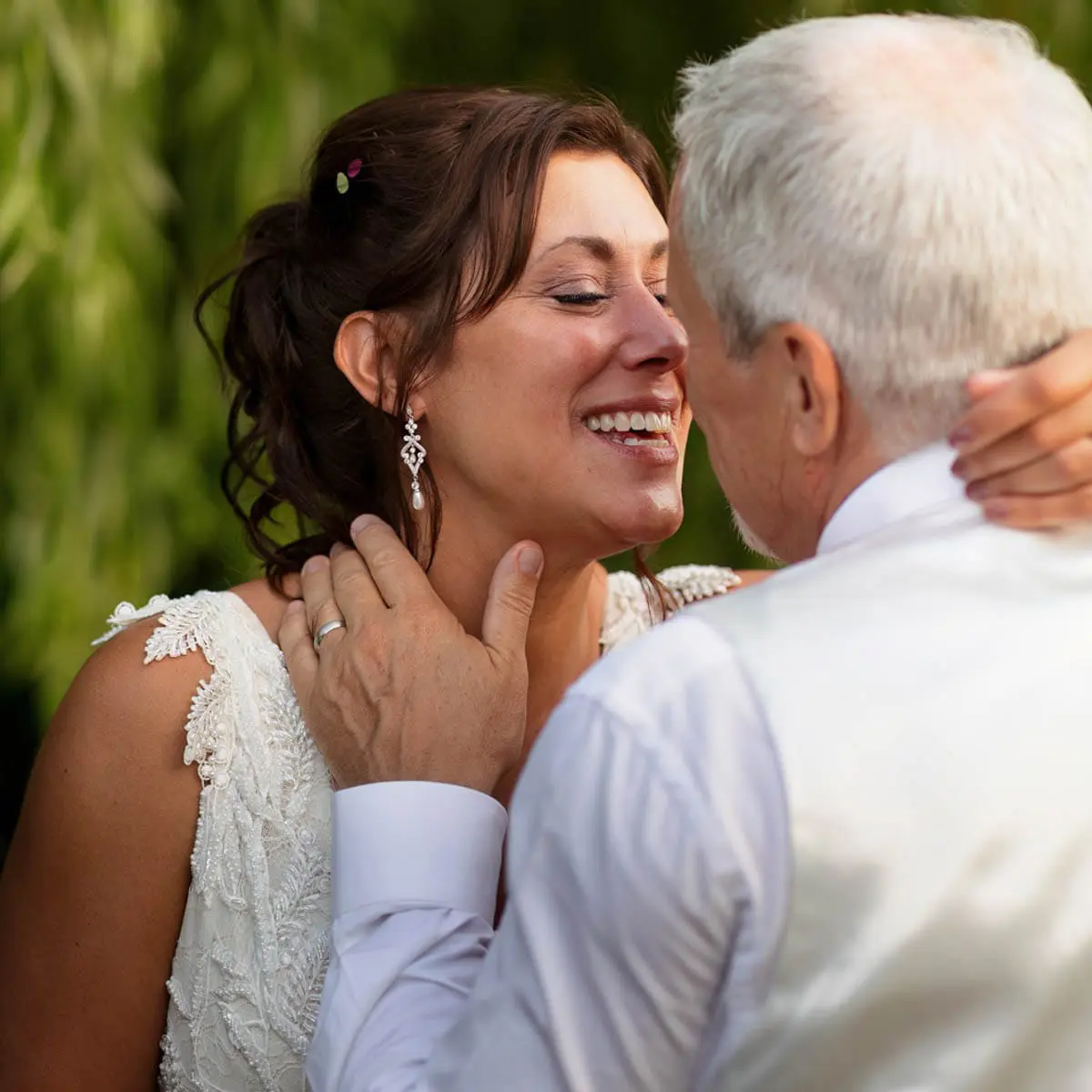 70_Gayle_and_Philip_Creative_Wedding_Photographer A bride and an older man, possibly her father, share an emotional embrace outdoors in a creative wedding photography style. Both are smiling warmly, with the bride wearing a beautiful white lace dress.